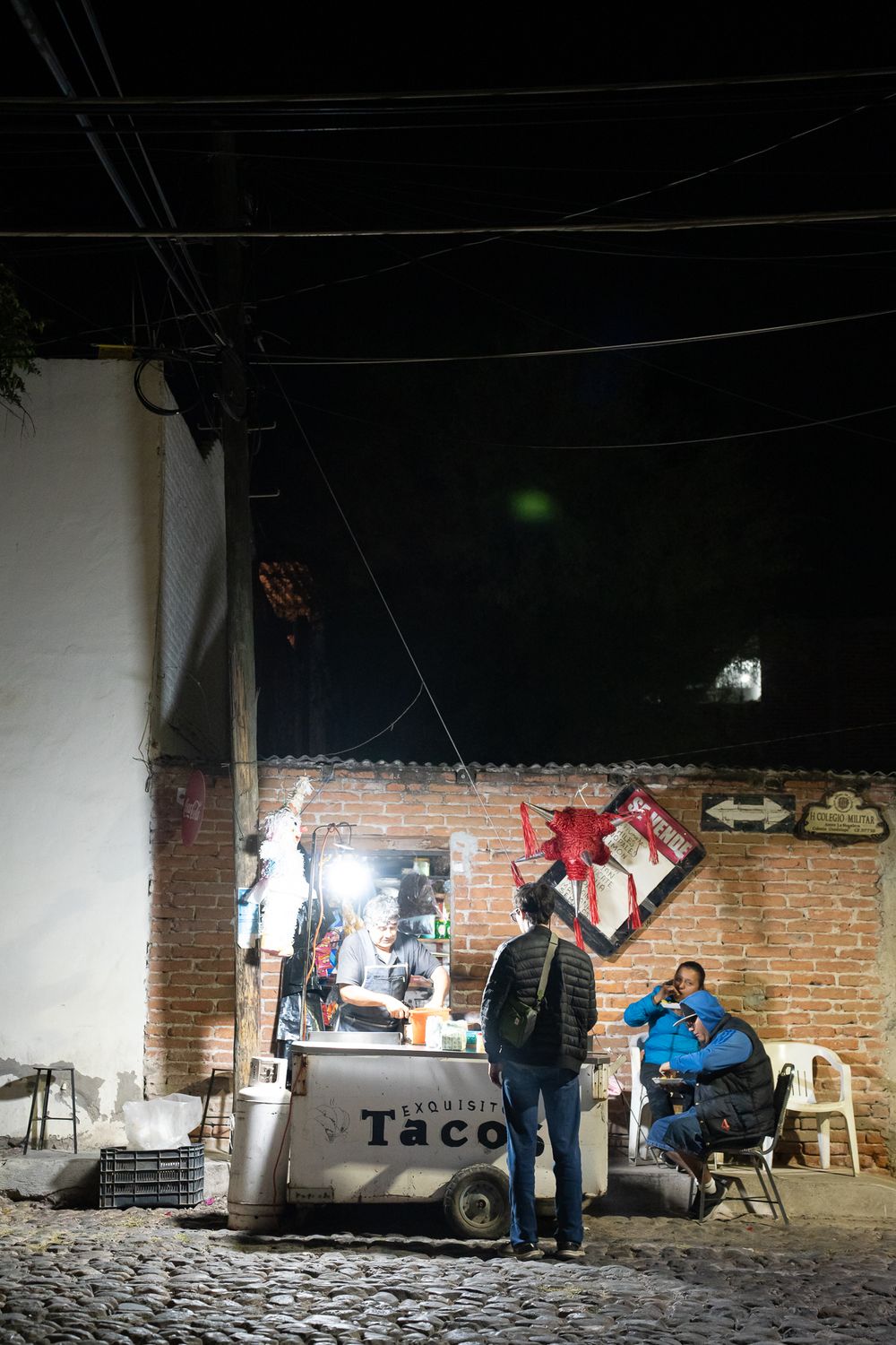 People gather at night in a dimly lit brick room with a taco stand setup.