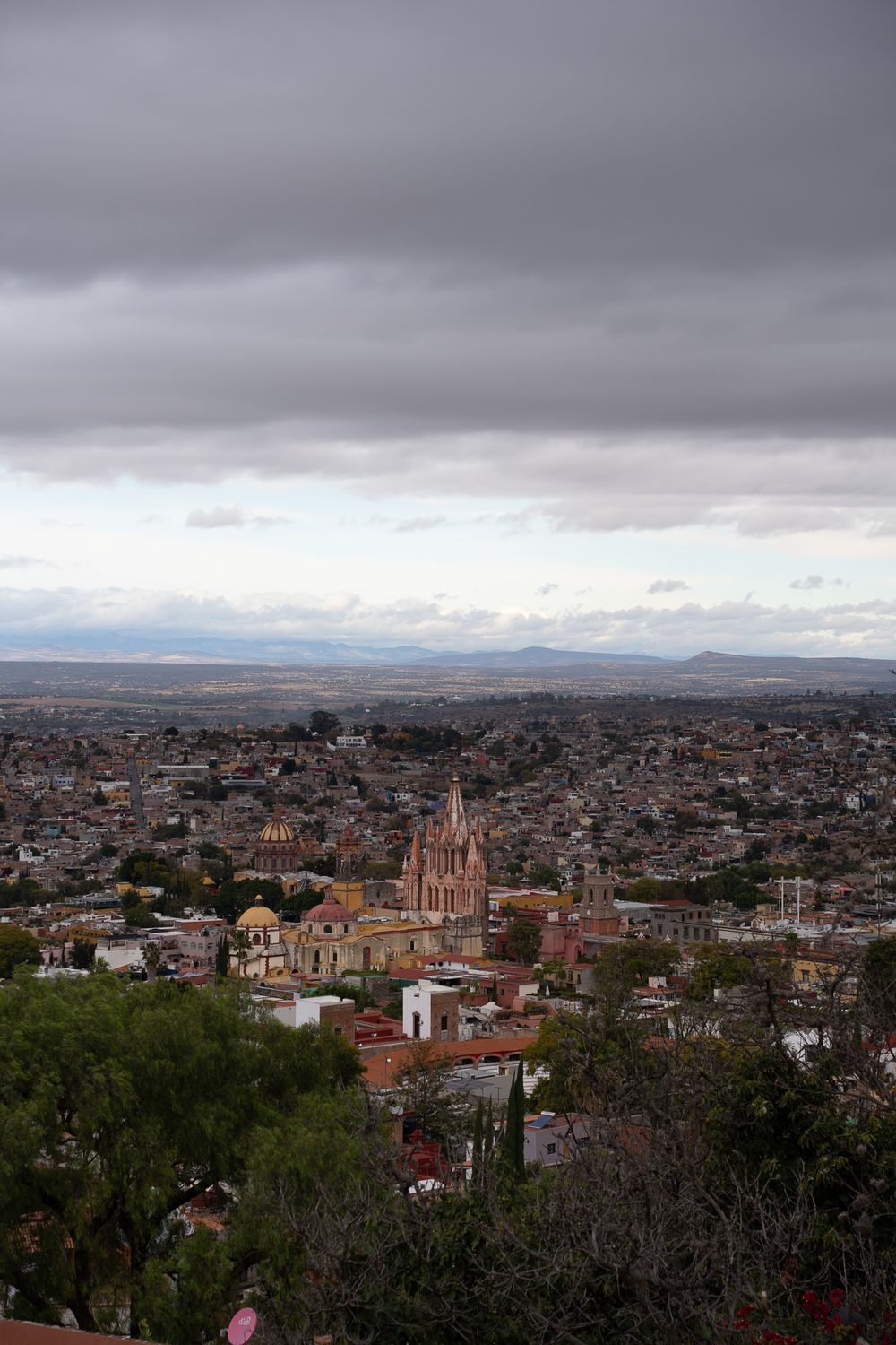 Panoramic view of San Miguel de Allende cityscape with historic church spires and buildings under cloudy skies.