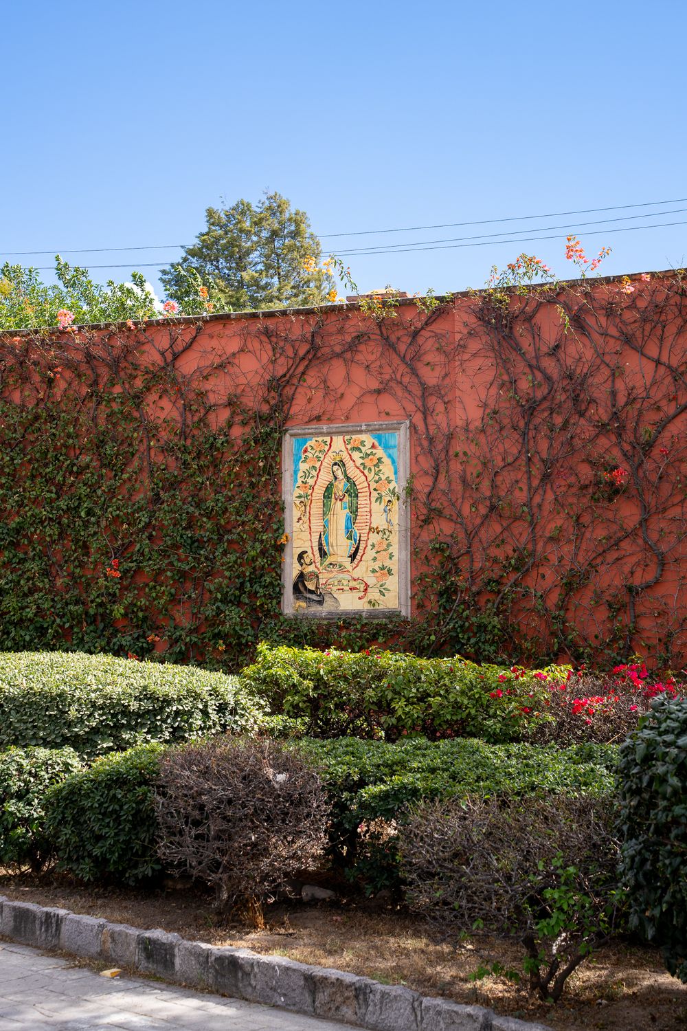 A religious mural on a red wall with decorative hedges and flowering plants along the base.