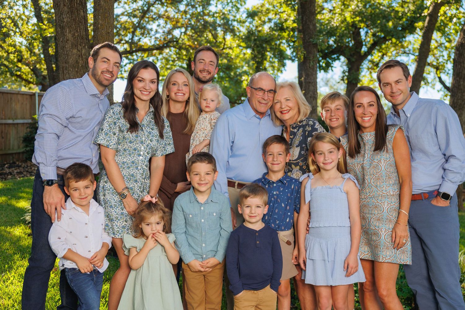 A large family group poses together outdoors in coordinated blue and gray outfits on a sunny summer day.