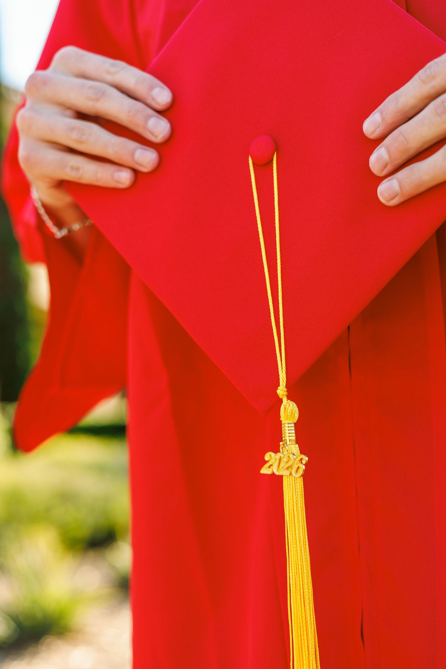 A yellow graduation tassel hangs against a bright red graduation gown with hands visible at the top.