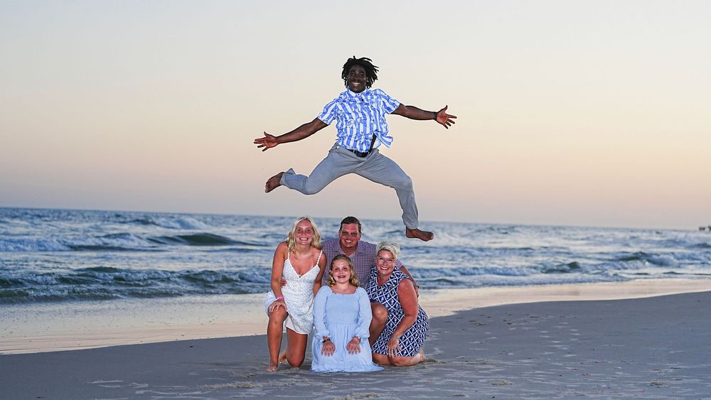 Family beach photo at sunset with four people posing together as a child joyfully leaps high in the air above them.