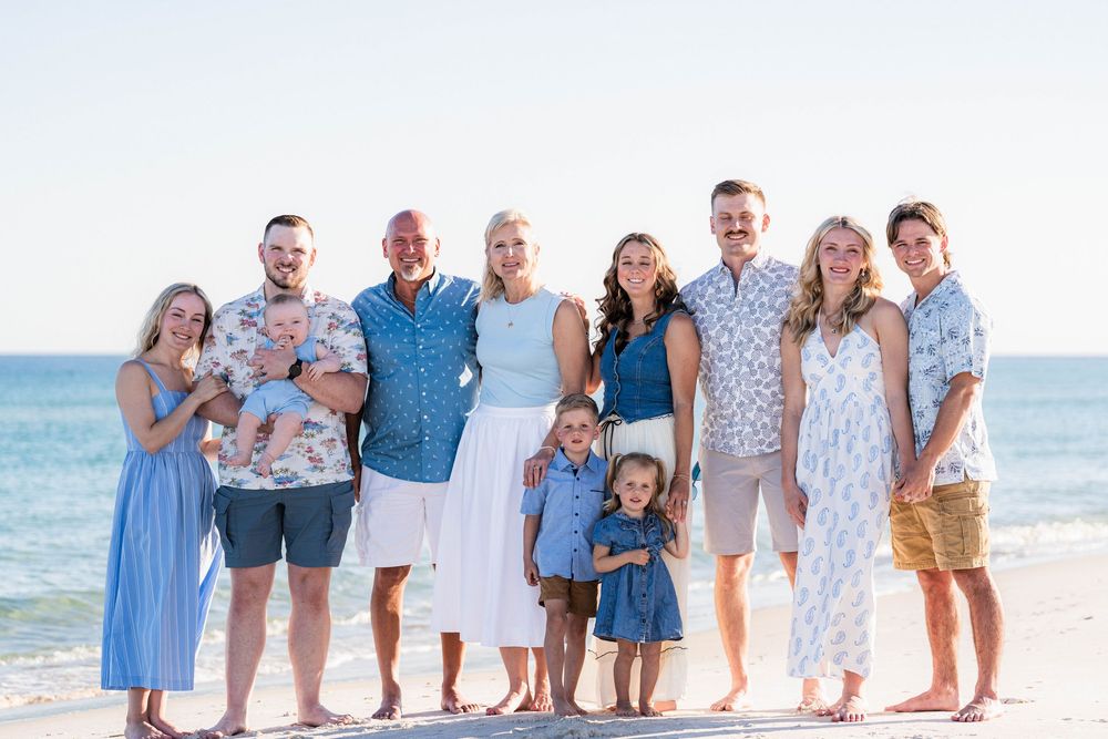 A joyful family of nine poses together on a sunny beach, dressed in coordinating blue and white outfits.