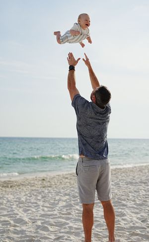 Father tossing laughing baby in the air on a sunny white sand beach with turquoise ocean waves in background.
