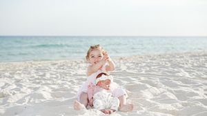 Two young children sit on a white sandy beach with calm blue ocean waves in the background on a sunny day.