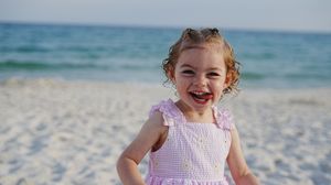 A cheerful toddler girl in a pink dress smiles on a sandy beach with blue ocean waves in the background.