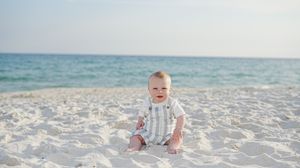 A happy baby sitting on white sand beach with calm blue ocean waves in the background on a sunny day.