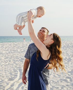 Happy couple lifting smiling baby overhead together on a bright sandy beach with ocean in the background.