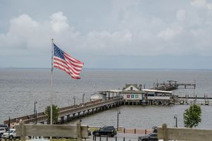 American flag waving over a waterfront pier with a small building and boats on a cloudy day.