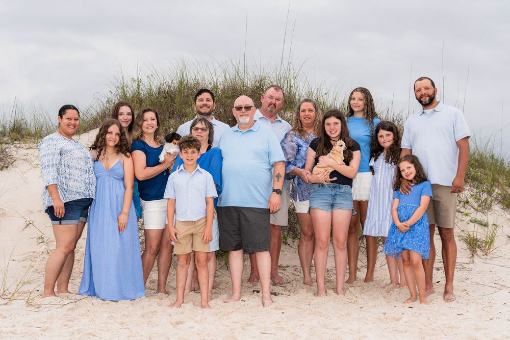 Large family group portrait on sandy beach with sea oats, wearing coordinated blue and white outfits.