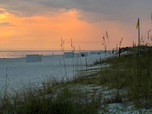 Coastal landscape at sunset with sea oats in foreground and orange sky reflecting on calm waters.