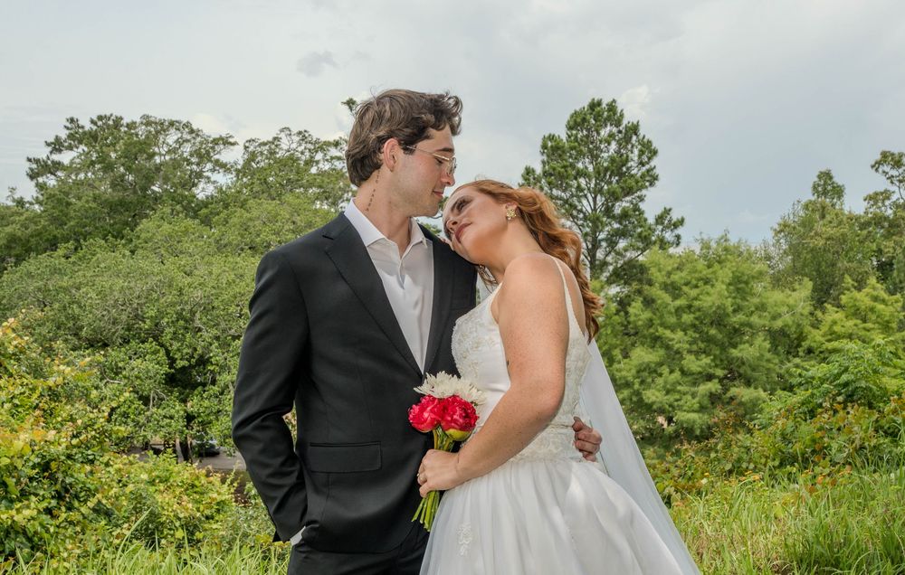 Bride and groom share a romantic moment outdoors, bride holding red bouquet in white gown, groom in black suit.