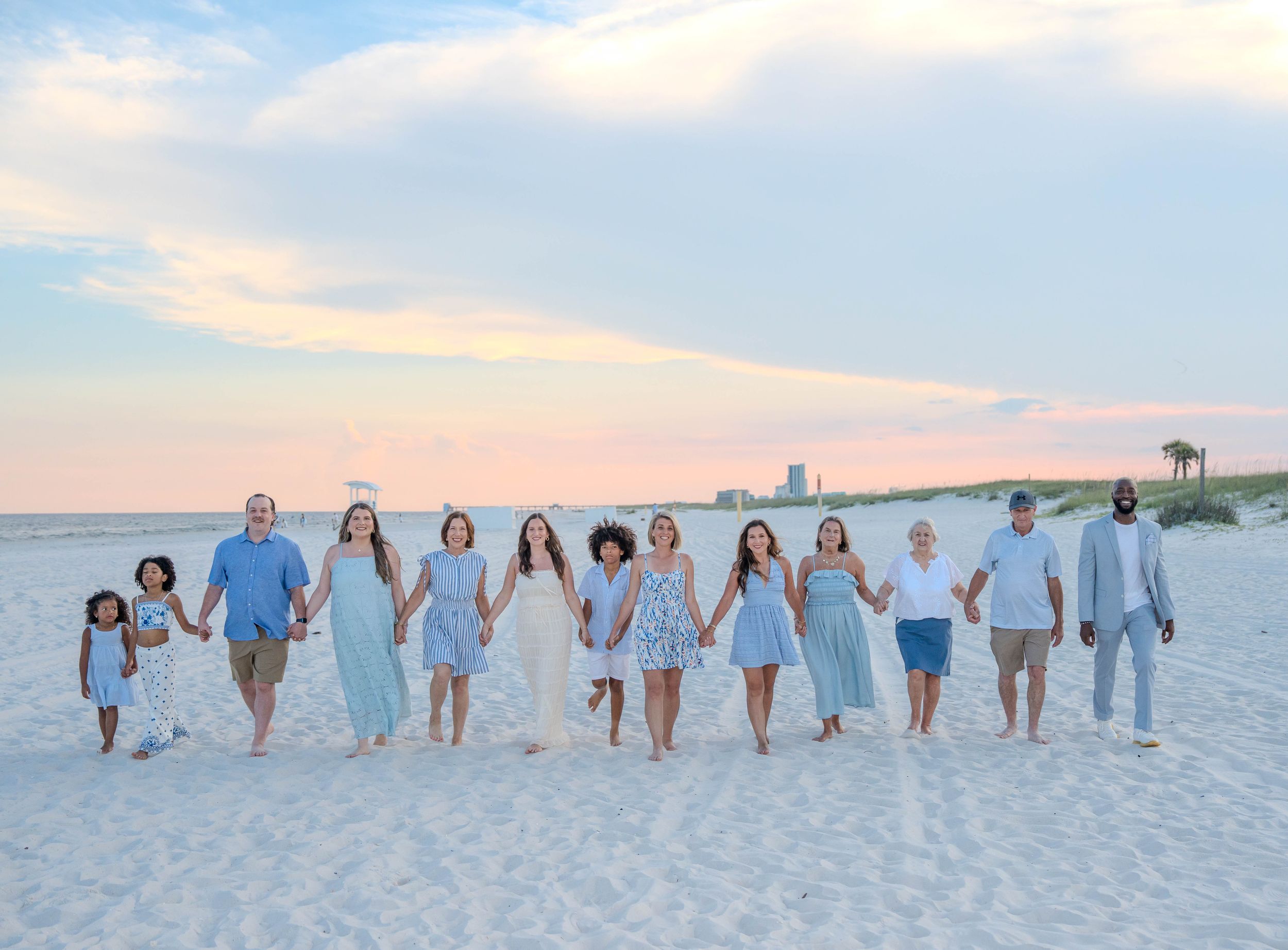 Large family group in coordinated blue outfits holding hands on a sandy beach at sunset.