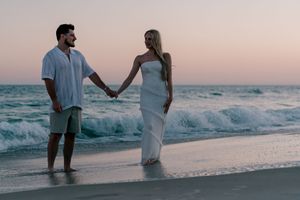 Couple holding hands on a sandy beach at sunset, man in white shirt and green shorts, woman in elegant white dress.