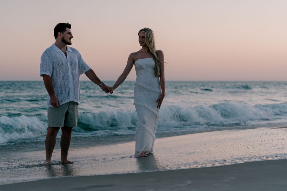 Couple holding hands on a sandy beach at sunset, man in white shirt and green shorts, woman in elegant white dress.
