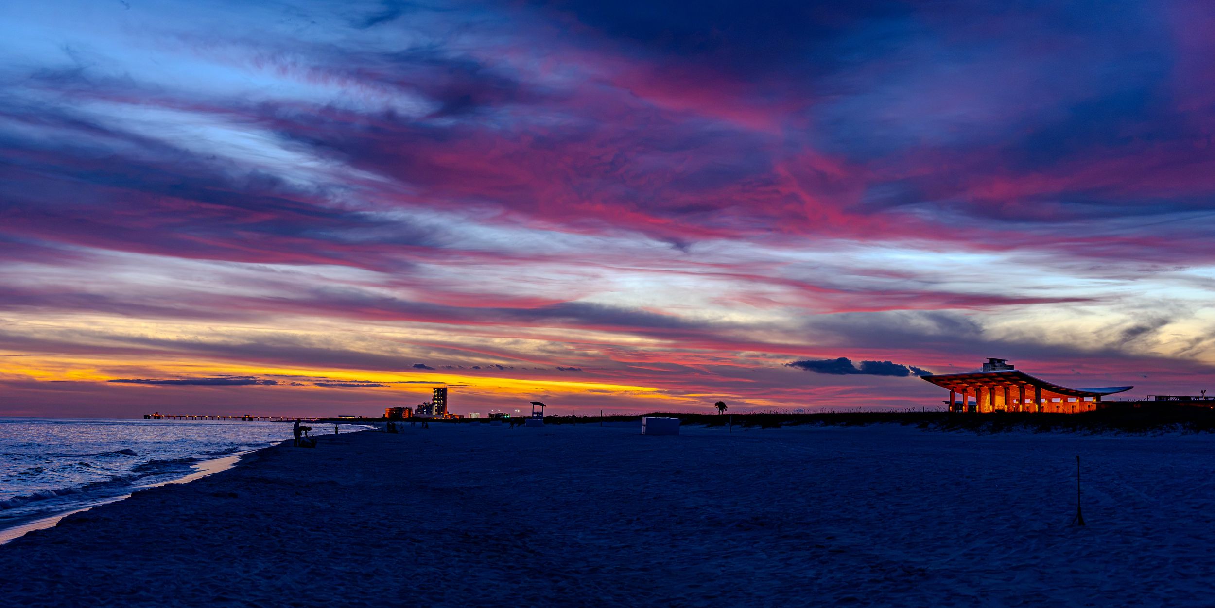 Stunning beach sunset with vibrant pink and purple clouds over calm ocean waters and illuminated pier at dusk.