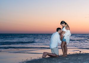 Couple sharing a romantic moment on a beach at sunset, man kneeling before woman by the ocean shoreline.