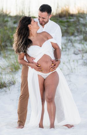 Couple sharing a romantic moment on white sand beach, woman in white maternity dress showing baby bump.