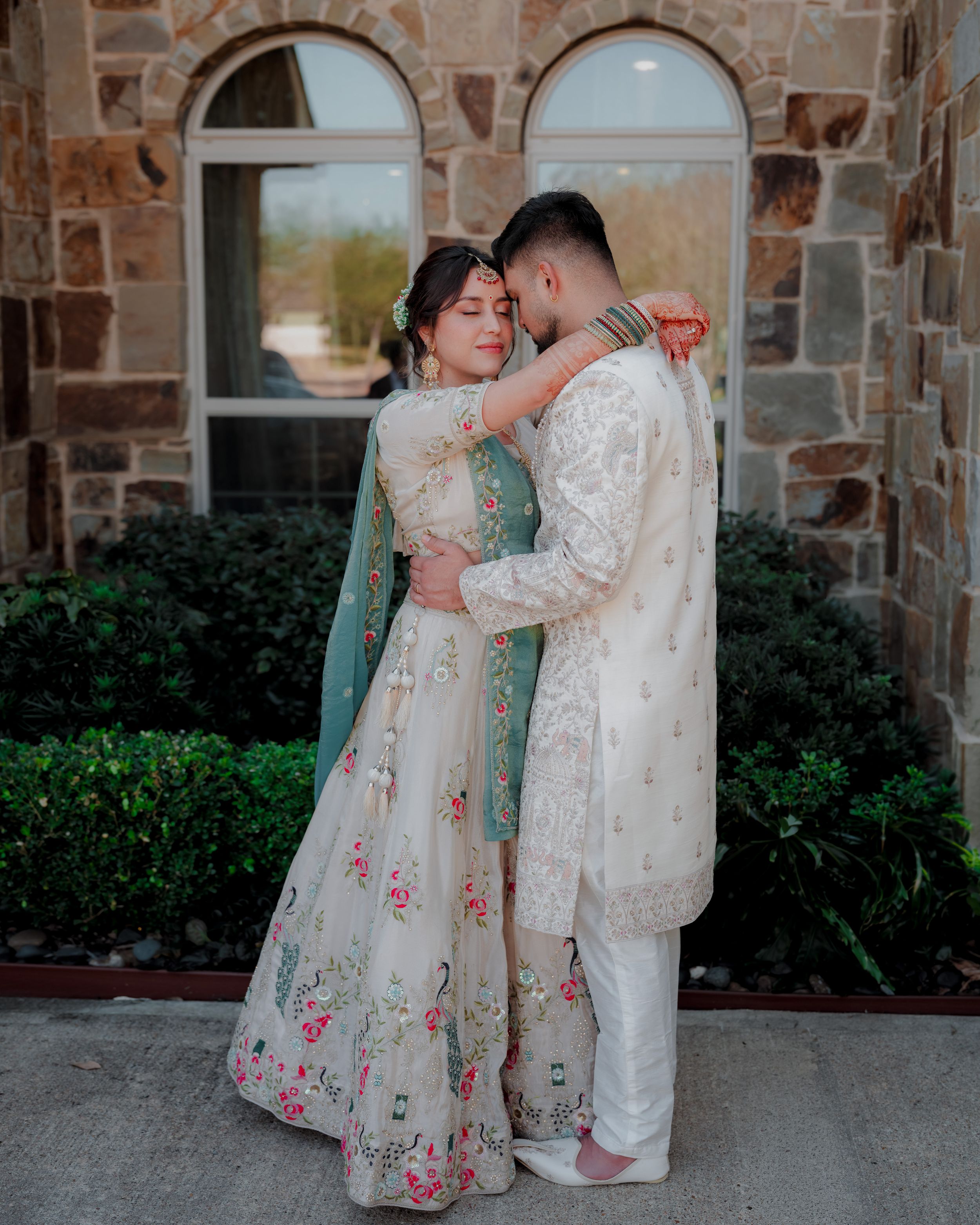 A couple in matching cream-colored traditional outfits embrace against a stone wall backdrop with greenery.