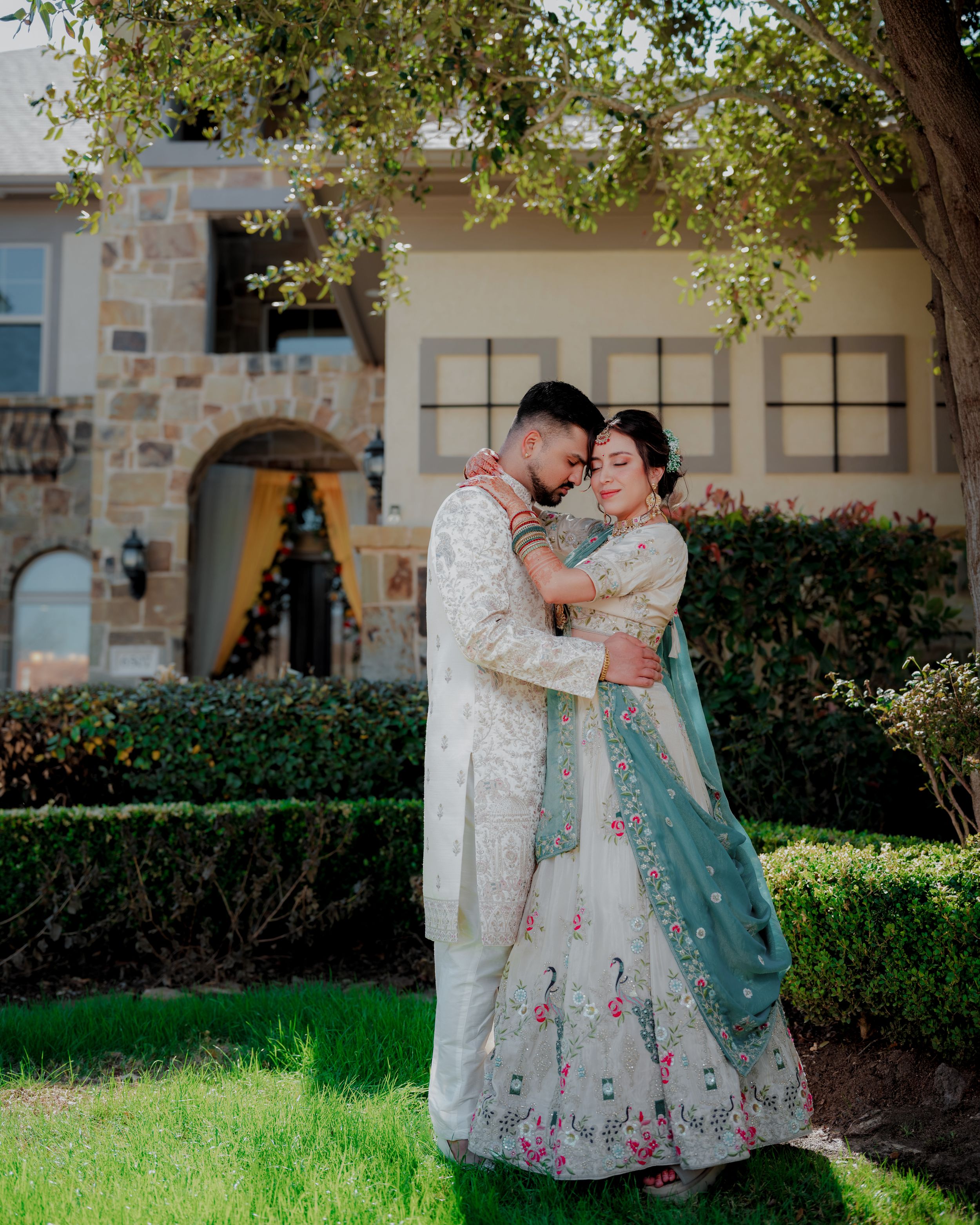 Two people in romantic formal attire embrace in a garden setting with stone buildings and greenery in the background.