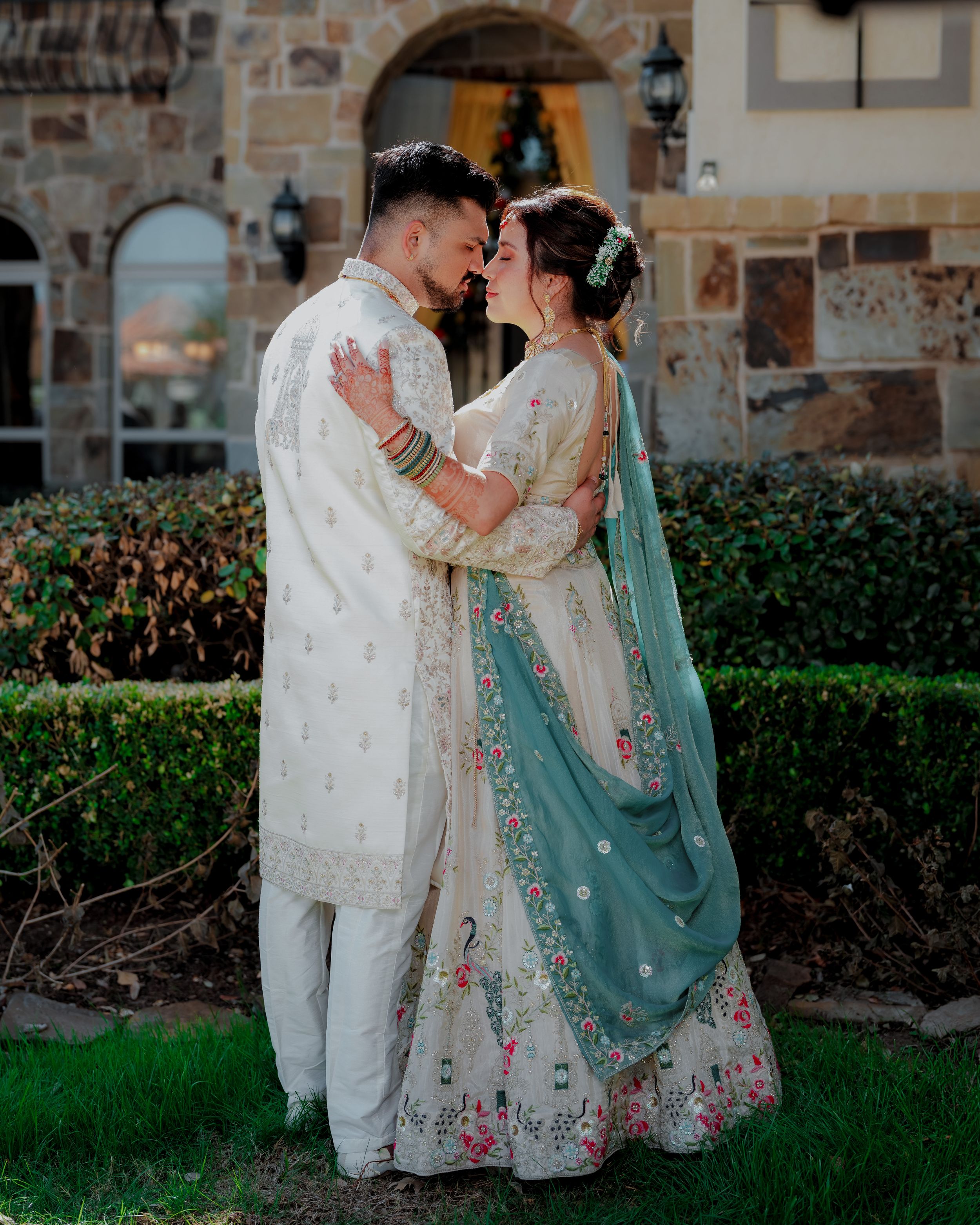 A couple in traditional South Asian wedding attire share intimate moments in a garden setting with stone walls.