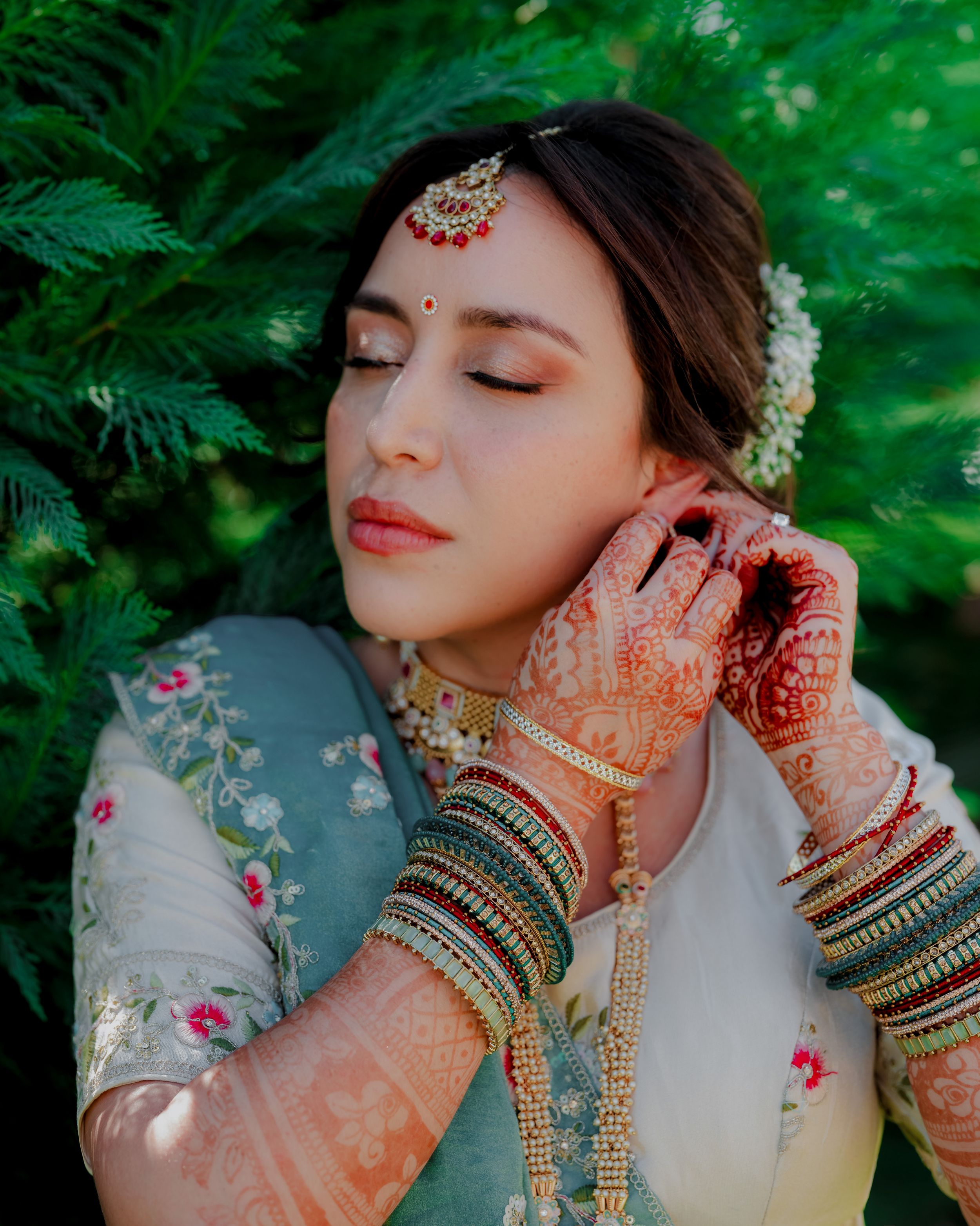 A bride in traditional Indian attire with henna adorned hands poses gracefully against lush green foliage.