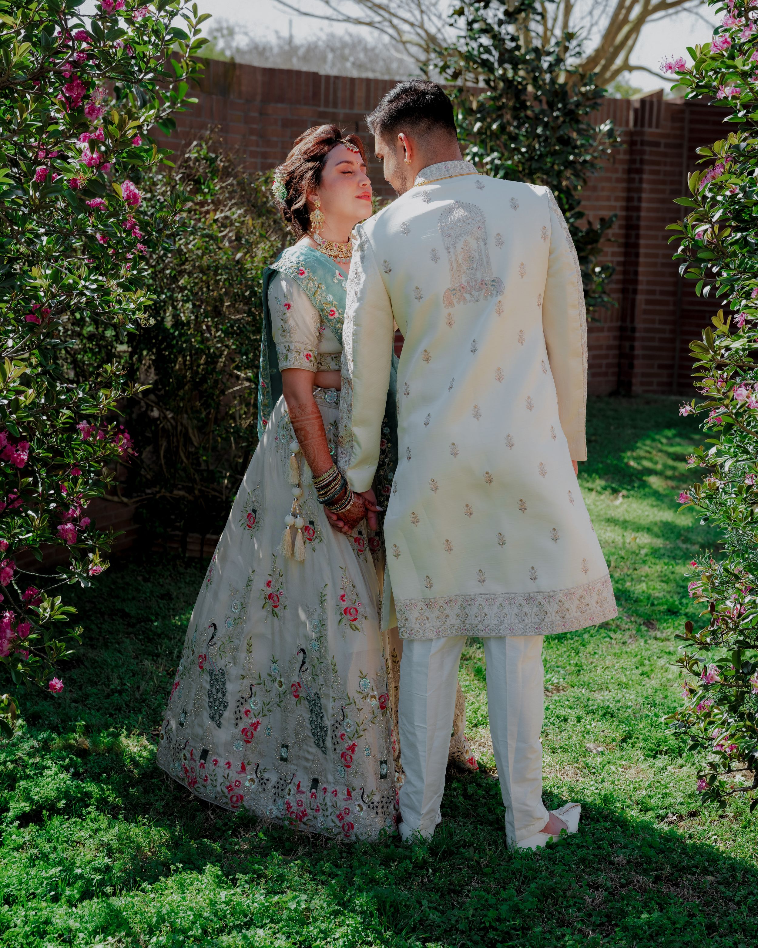A couple in traditional white attire poses together in a garden setting with blooming pink flowers and greenery.