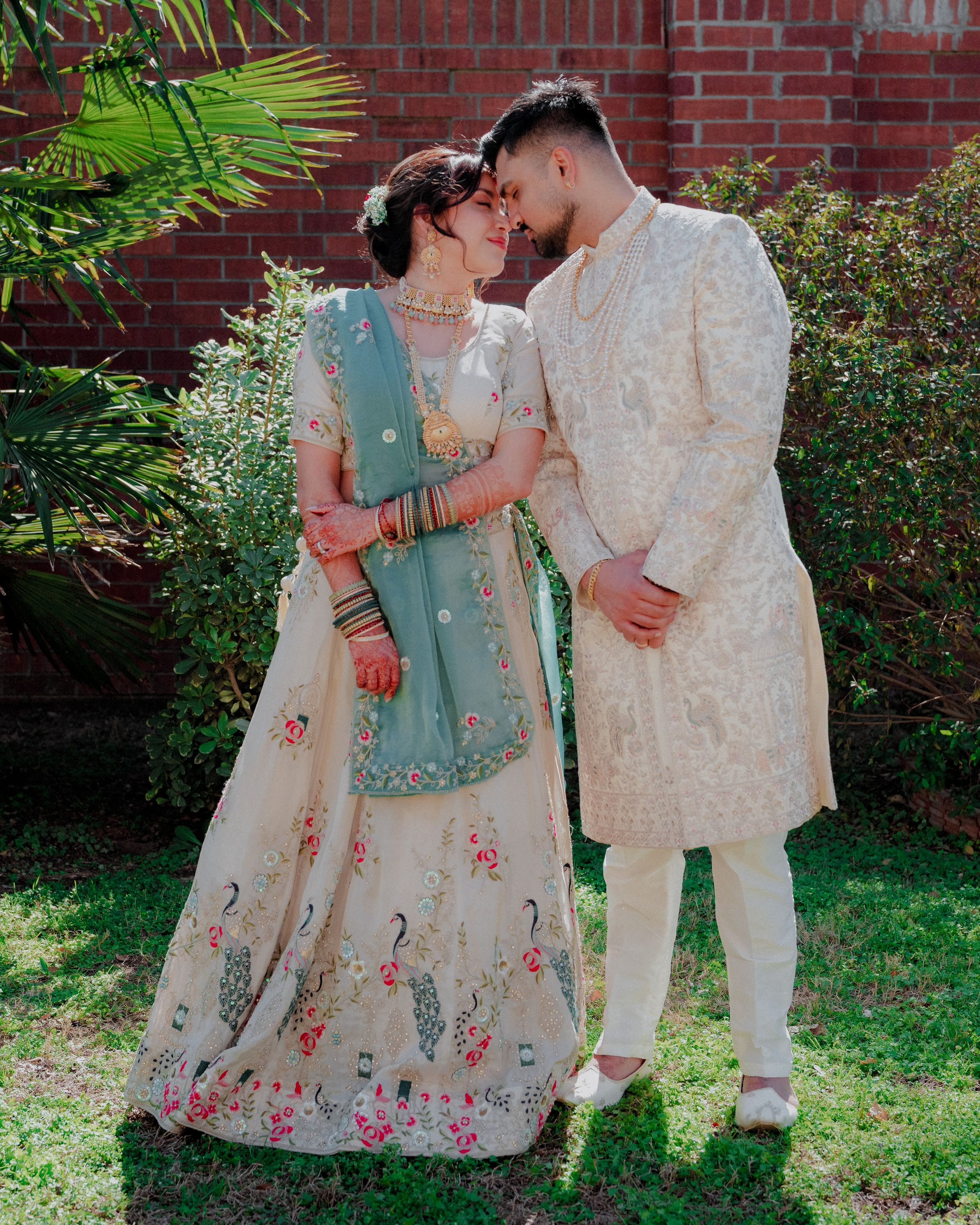 A couple in traditional South Asian wedding attire share an intimate moment against a brick wall and garden backdrop.