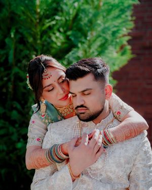 A romantic couple in traditional South Asian wedding attire embrace in front of lush greenery.