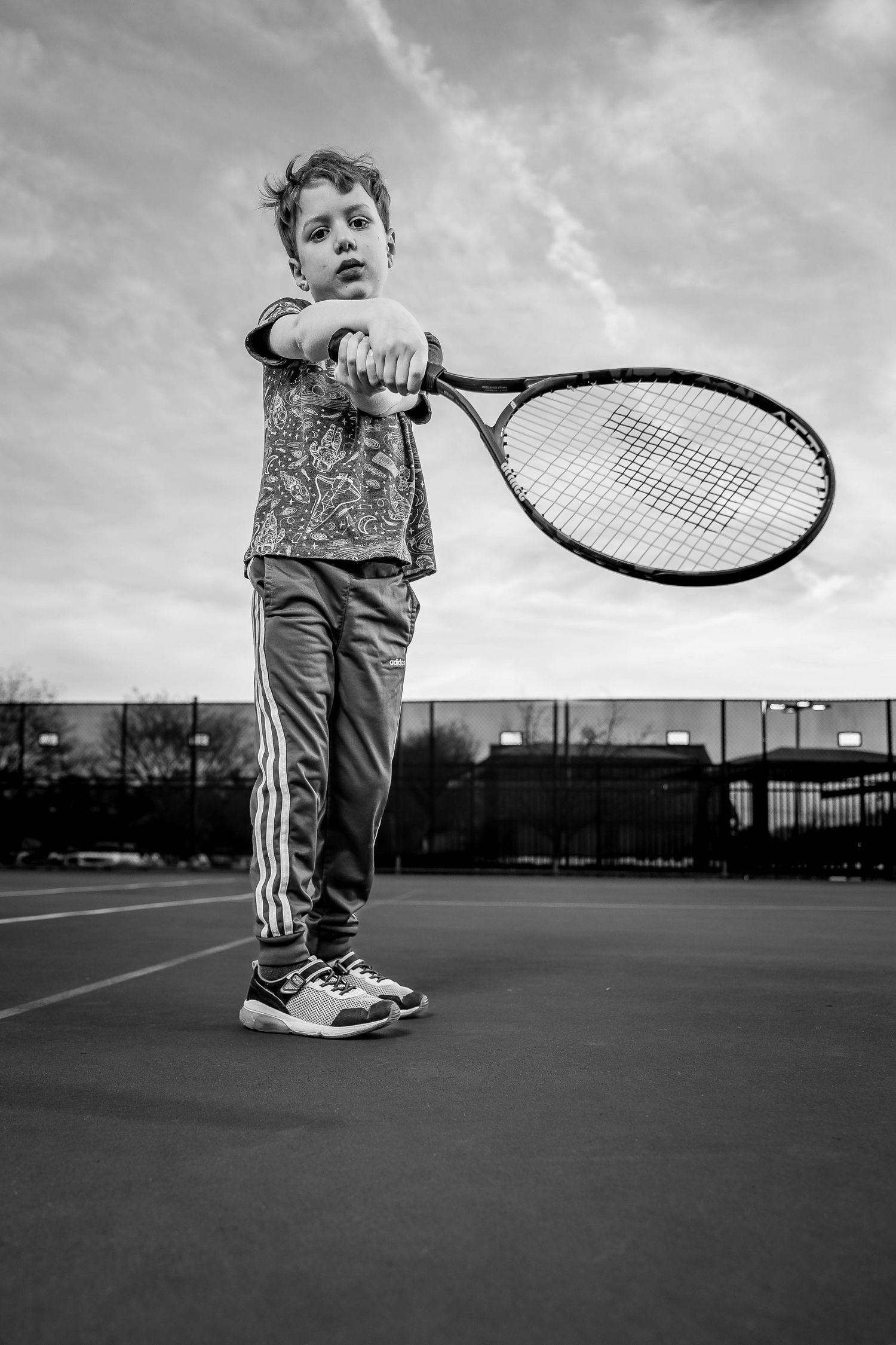 A young tennis player in striped pants holds a large racquet on an outdoor court in dramatic black and white.