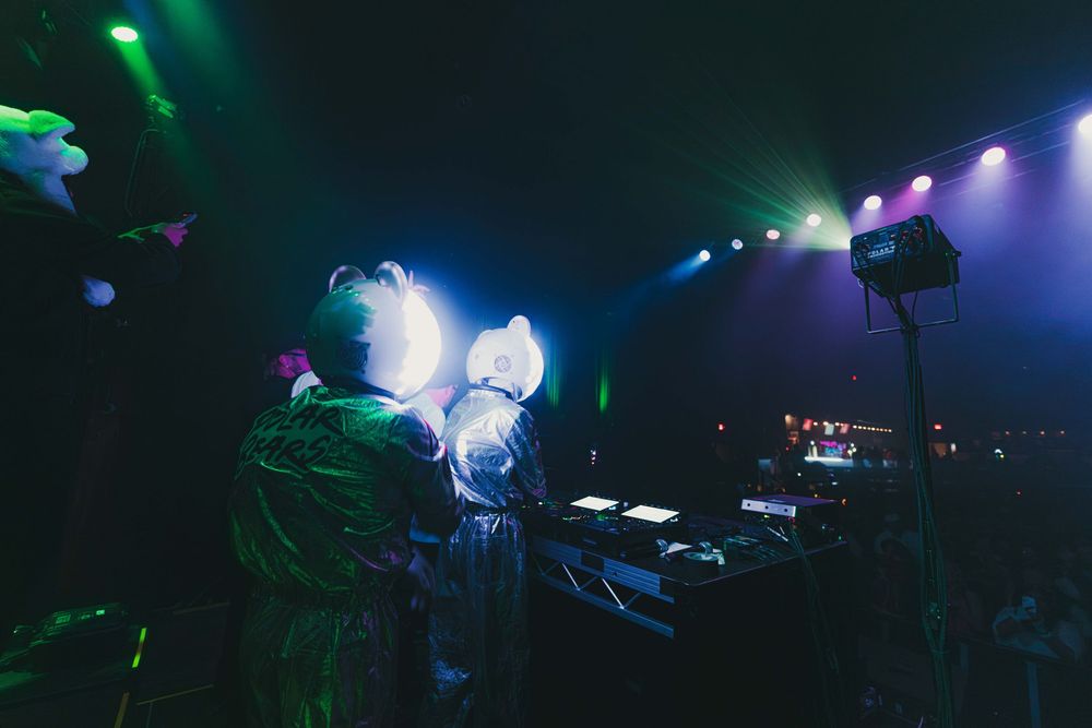 Silhouettes of concert performers on stage with dramatic green and purple lighting effects in a dark venue.