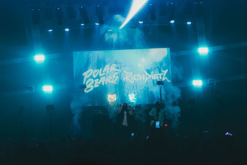 Blue stage lights create a moody atmosphere during a live performance with silhouettes visible against the dark background.