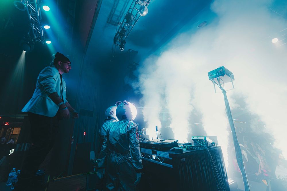 Musicians performing on a foggy stage illuminated by dramatic turquoise spotlights during a live show.