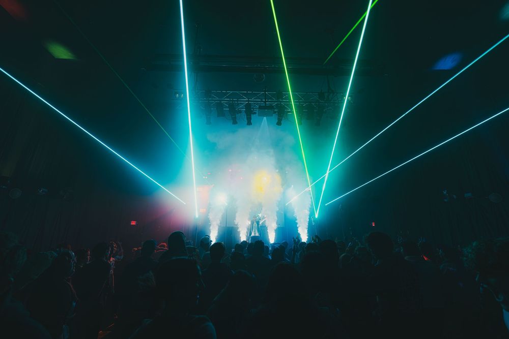 Concert stage illuminated with vibrant blue and green laser beams creating a dramatic atmosphere over silhouetted crowd.