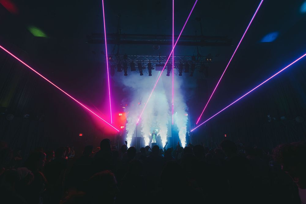 Colorful laser beams illuminate a dark concert venue with vibrant pink, blue and purple lights creating a dramatic atmosphere.
