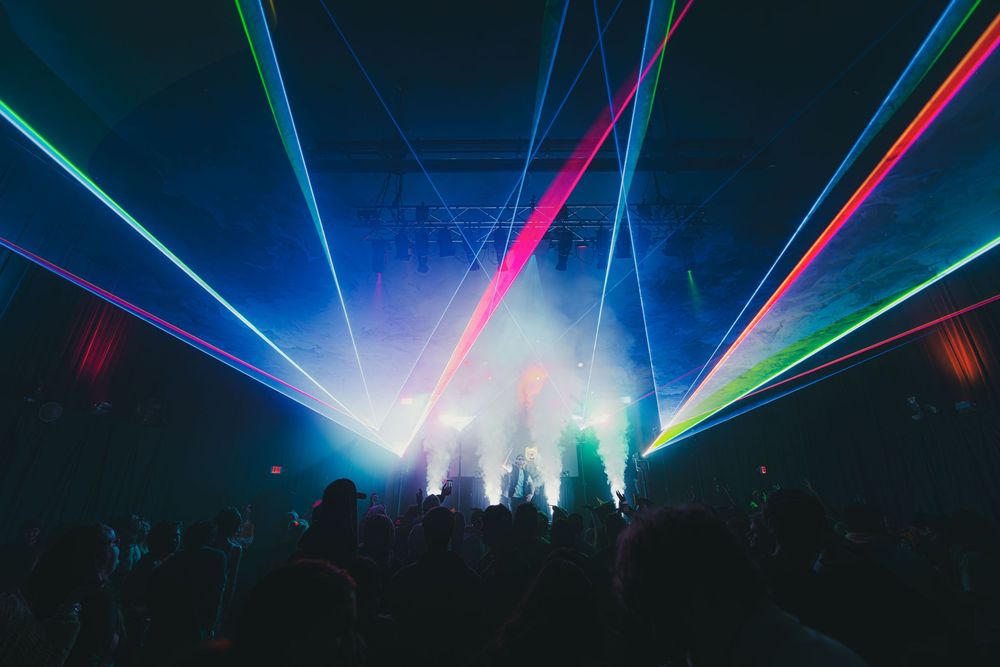 Colorful laser beams illuminate a dark concert venue with blue, pink and green lights creating a dramatic atmosphere.