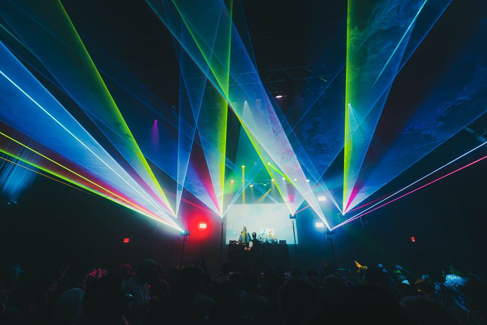 Colorful laser beams illuminate a dark concert venue with blue and green lights creating a dramatic nightclub atmosphere.
