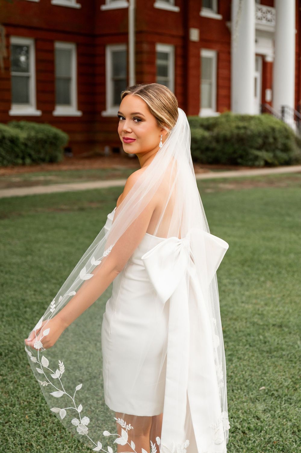 Elegant bride in white dress with flowing veil poses on manicured lawn.