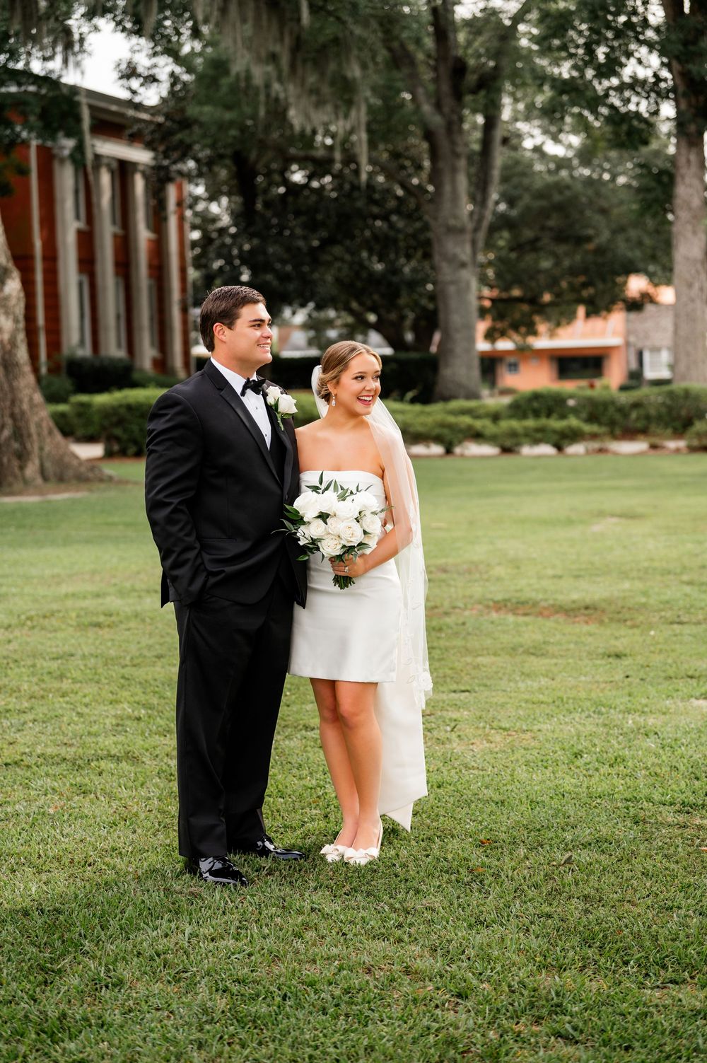 Wedding couple takes creative photos on grassy lawn near brick building.