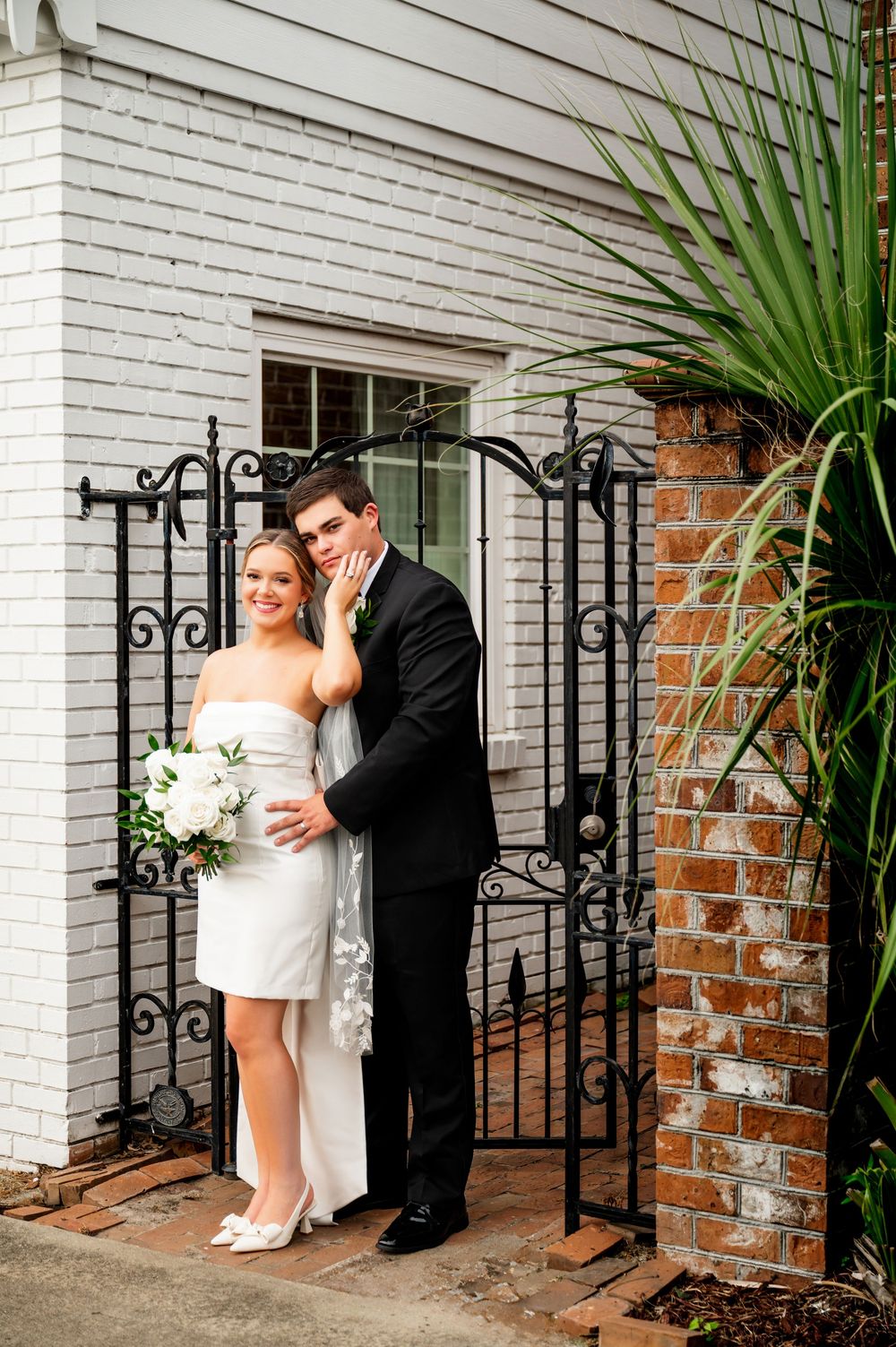 Wedding couple poses by decorative iron gate and white brick wall.