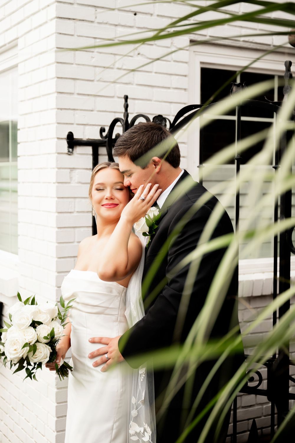 Romantic wedding photos through palm fronds against white brick backdrop.
