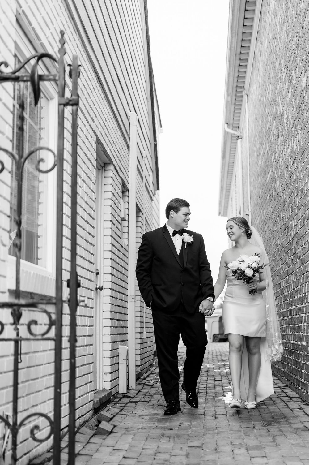 A couple in wedding attire walks together down a narrow brick alleyway between buildings.