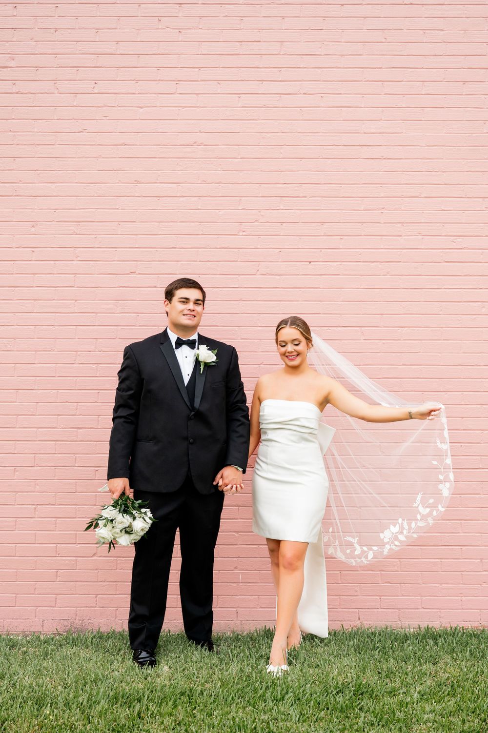Wedding couple poses against a pink brick wall with the bride holding her veil.