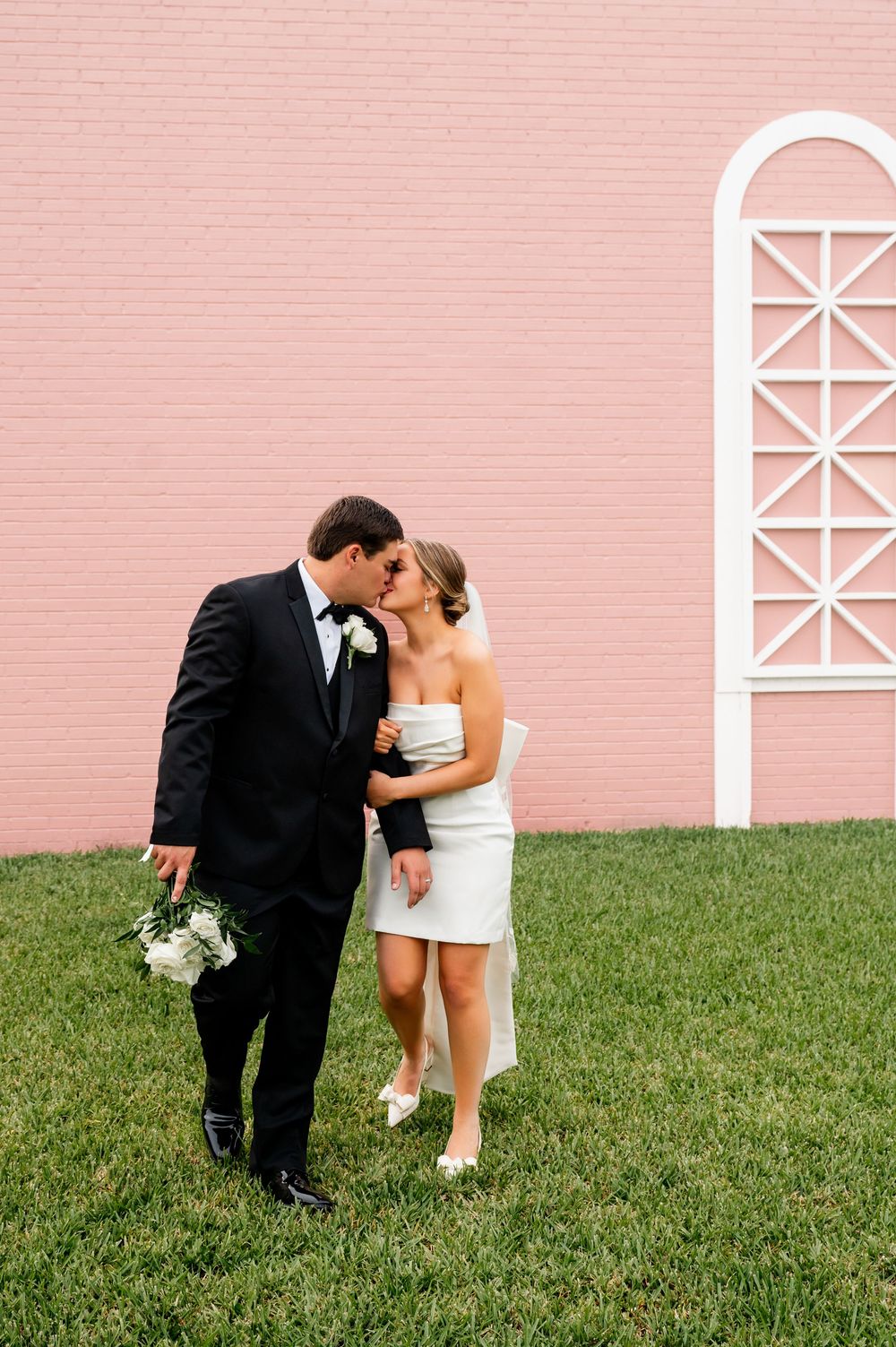 Newlyweds share an intimate moment against a pink wall with white architectural details.