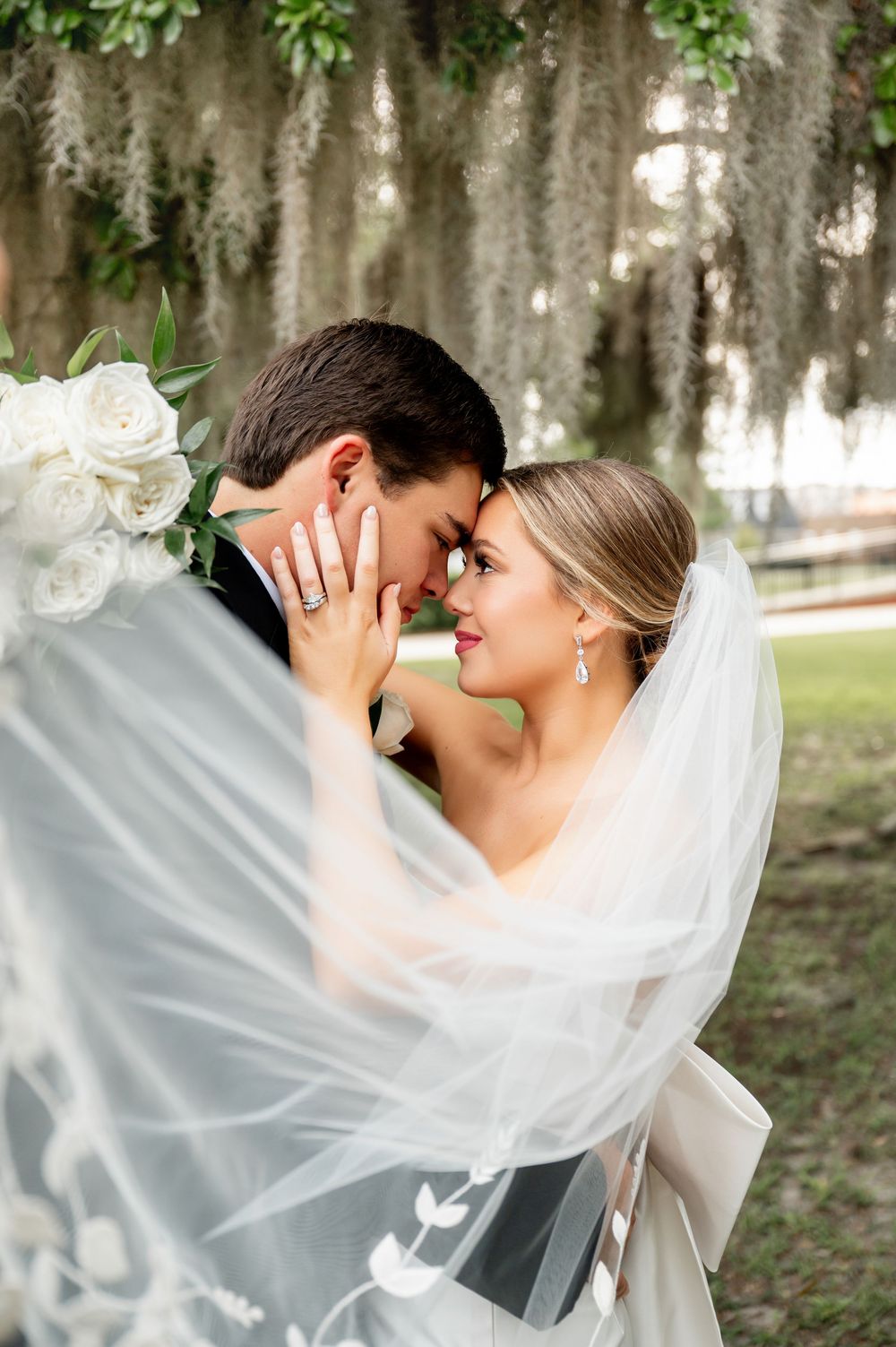 Intimate moment between newlyweds captured through flowing veil under Spanish moss.