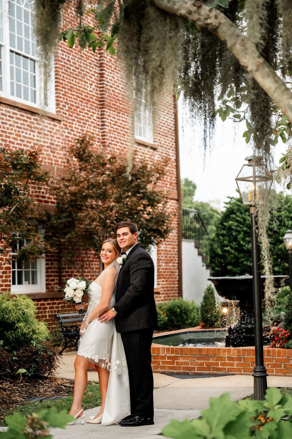 Wedding couple poses by historic brick building with Spanish moss hanging overhead.
