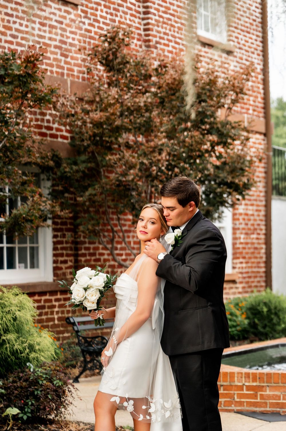Elegant wedding portrait against brick building with landscaped garden.