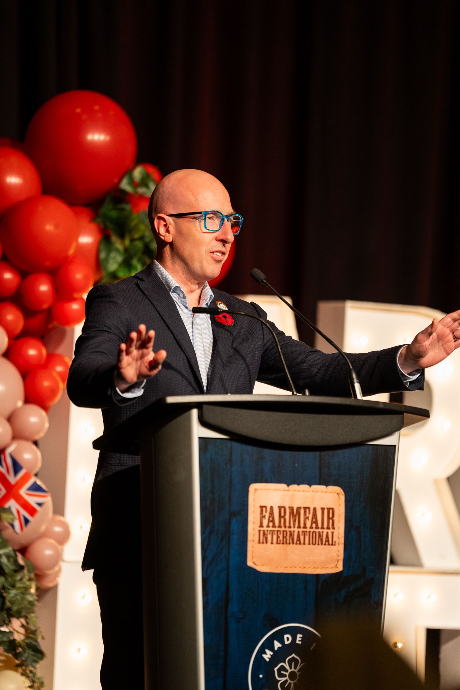 Bald man in suit speaks at Farmfair International podium with red and white balloon decorations at an indoor event.