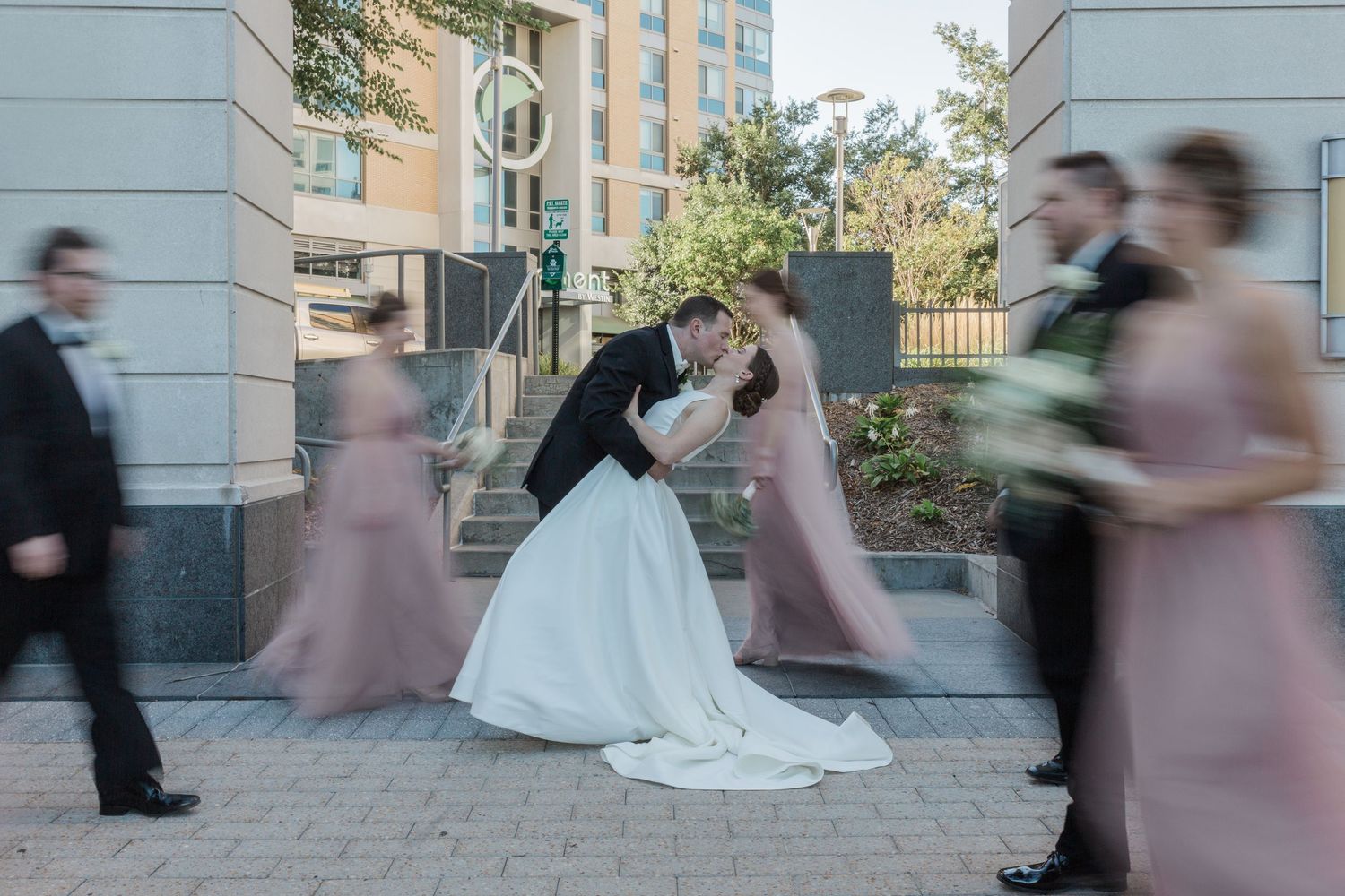 Wedding party members rush past in a motion blur effect while the bride and groom remain still for photos.