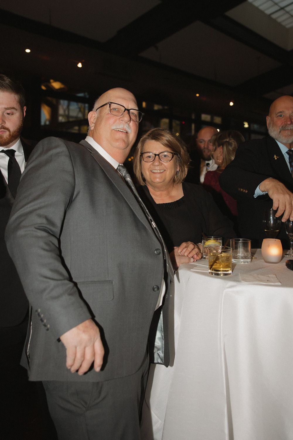 Guests in formal attire mingle at an elegant evening reception with white tablecloths and ambient lighting.